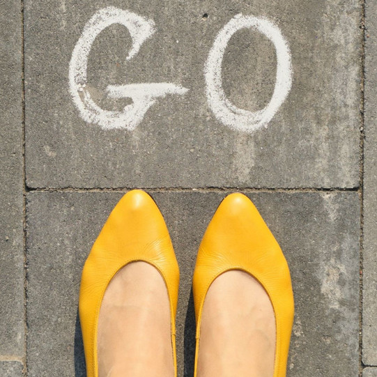 Foto: Frauenfüße in gelben Ballerinas, auf Pflastersteine, vor den Füßen steht in Kreide das Wort "Go".