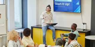 Ein Mann präsentiert vor einer Gruppe an einem Konferenztisch; auf einem Bildschirm hinter ihm steht ‚Start it up!‘.