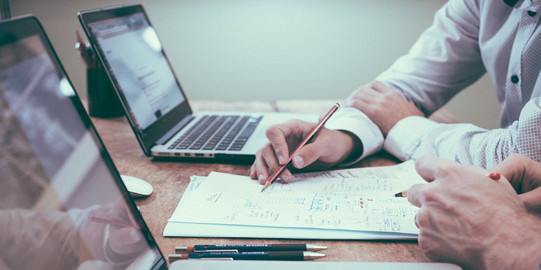 Zwei Laptop auf einem Schreibtisch und zwei Menschen arbeiten über einem Blatt Papier.