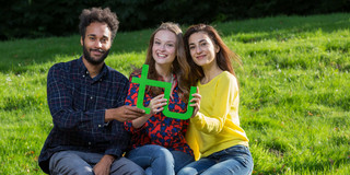 Studierende mit TU-Logo Drei Studierende sitzen auf einer Bank und halten gemeinsam ein grünes TU-Logo in den Händen.