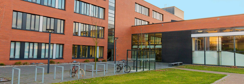 Seminarraumgebäude Seminarraumgebäude: Ein rotes dreistöckiges Gebäude mit schwarz gerahmten Fenstern, von der Seite fotografiert. Davor eine Wiese und Fahrradständer sowie Straßenlaternen.
