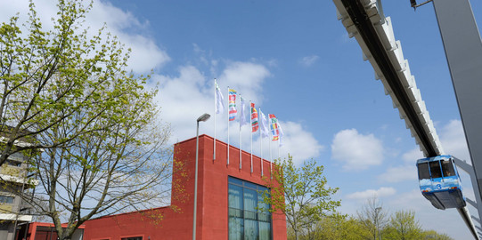 Internationales Begegnungszentrum mit H-Bahn Von Bäumen umgebenes Gebäude des Internationalen Begegnungszentrums und im Hintergrund blauer Himmel mit wenigen Wolken. Rechts im Bild die H-Bahn.