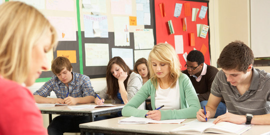 Unterricht im Klassenraum Schülerinnen und Schüler sitzen in einer Klasse und schreiben in Hefte, vor ihnen sitzt eine Lehrerin an einem Pult.
